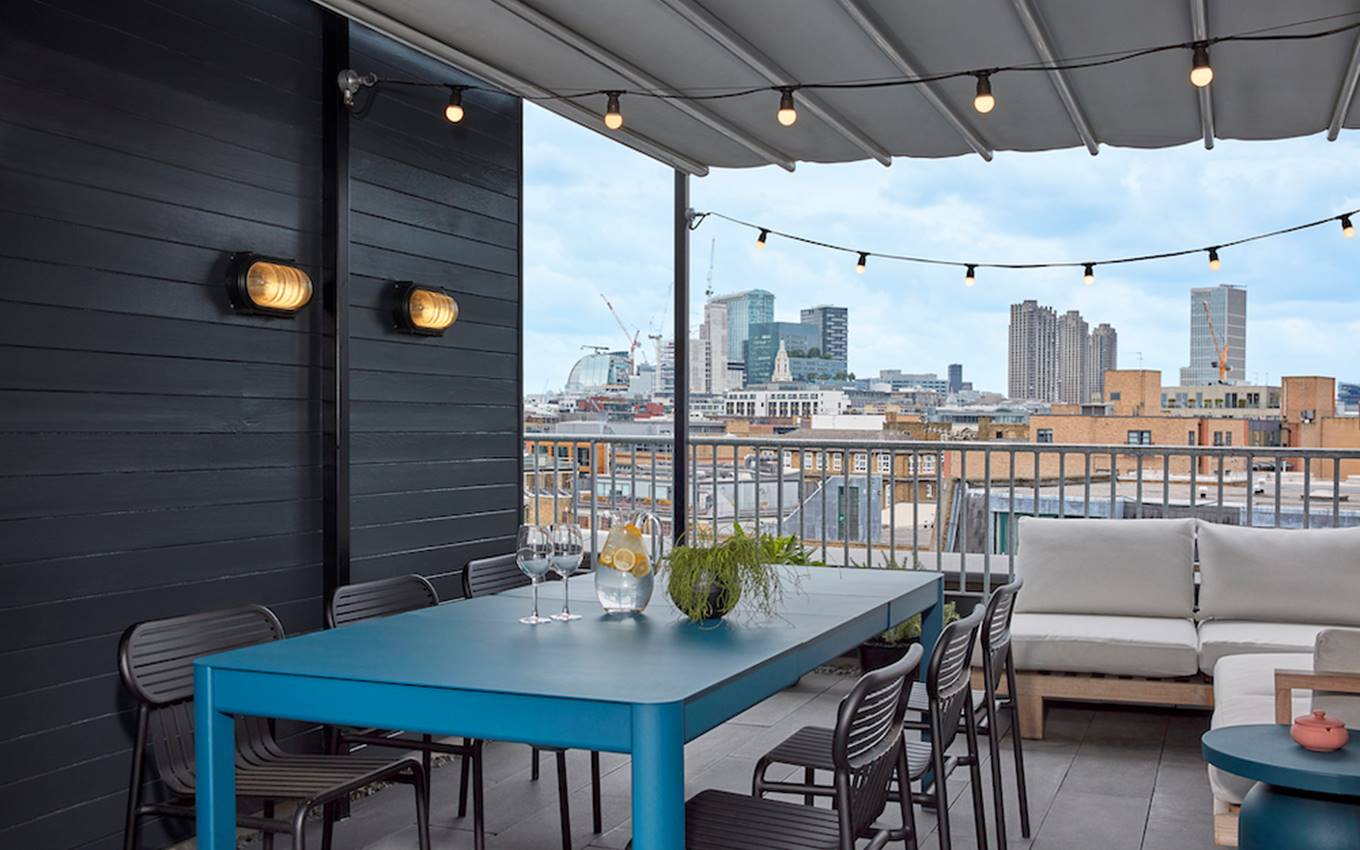 A rooftop terrace at One Hundred Shoreditch with a blue table set with glasses, offering London views under string lights.