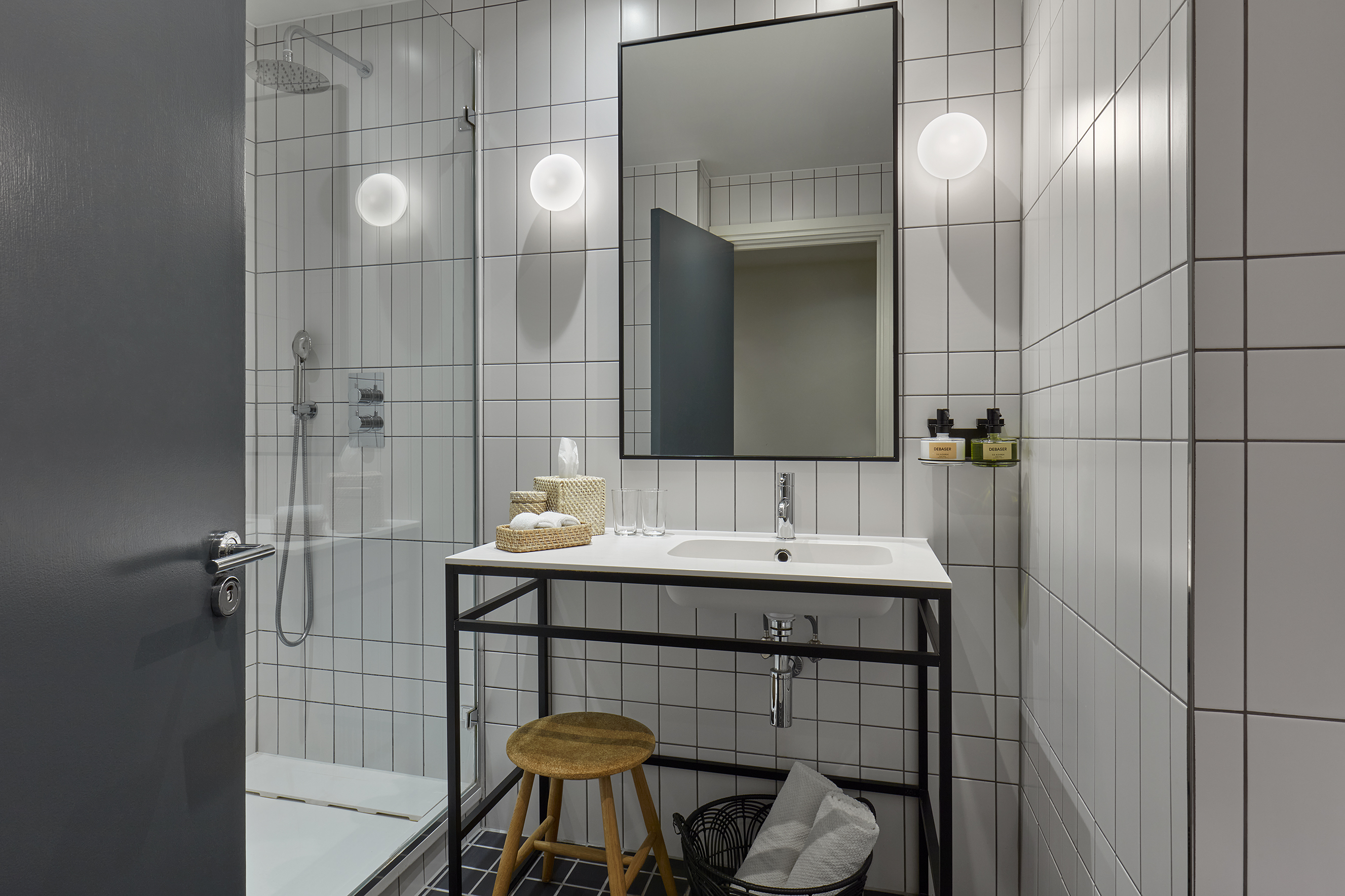 Minimalist bathroom at a London hotel with white tiles, modern sink on a black frame, and a small wooden stool.