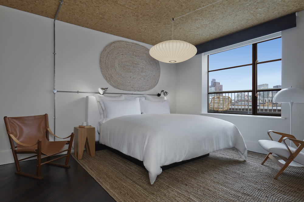 Minimalist hotel room in London with a white bed, woven wall art, and a large window showcasing a view of the city.