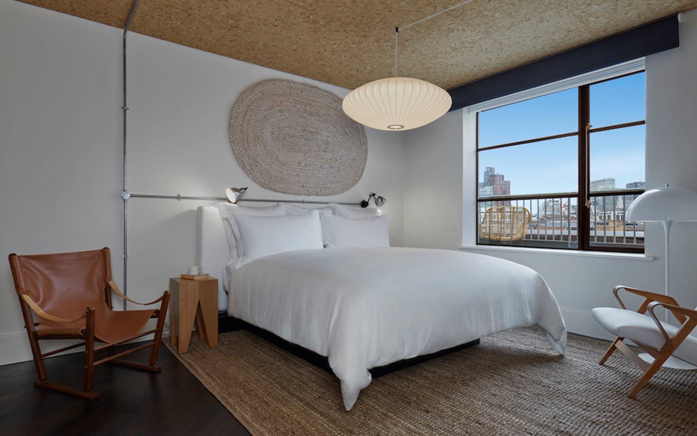 Minimalist hotel room in London with a white bed, woven wall art, and a large window showcasing a view of the city.