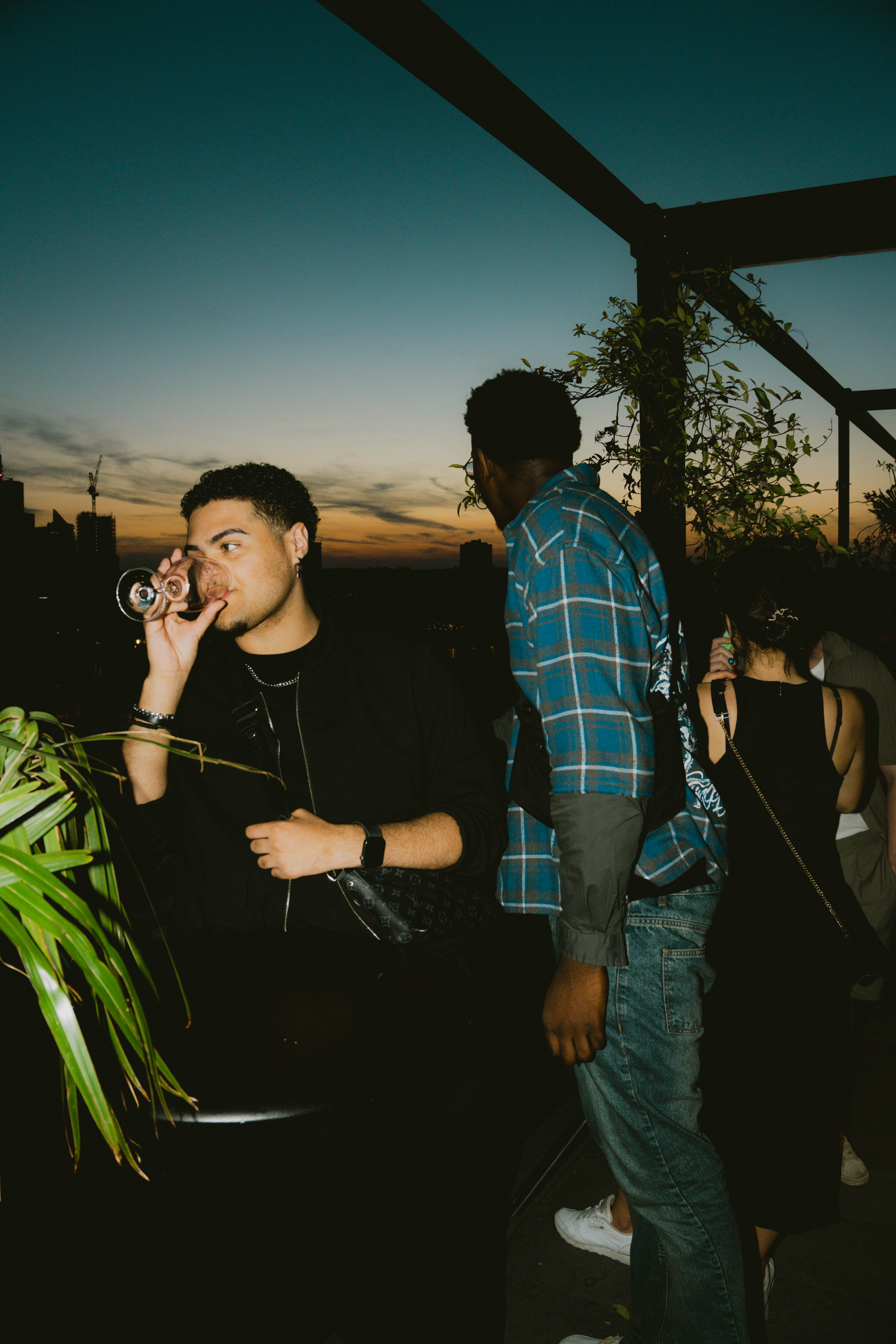 Man in black outfit sipping wine at a rooftop party, with a sunset sky and guests in the background at Kaso in London.