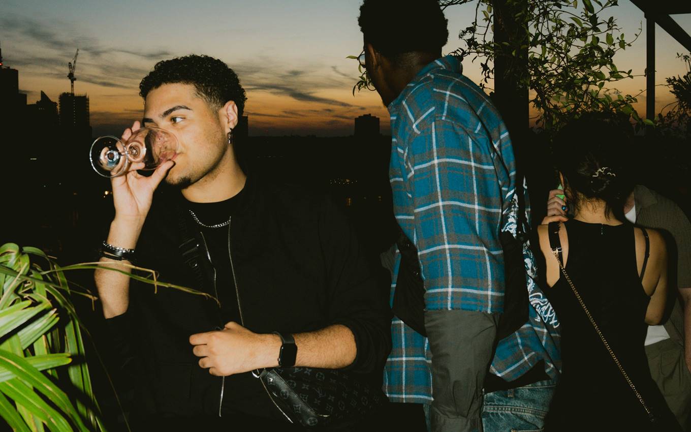 Man in black outfit sipping wine at a rooftop party, with a sunset sky and guests in the background at Kaso in London.