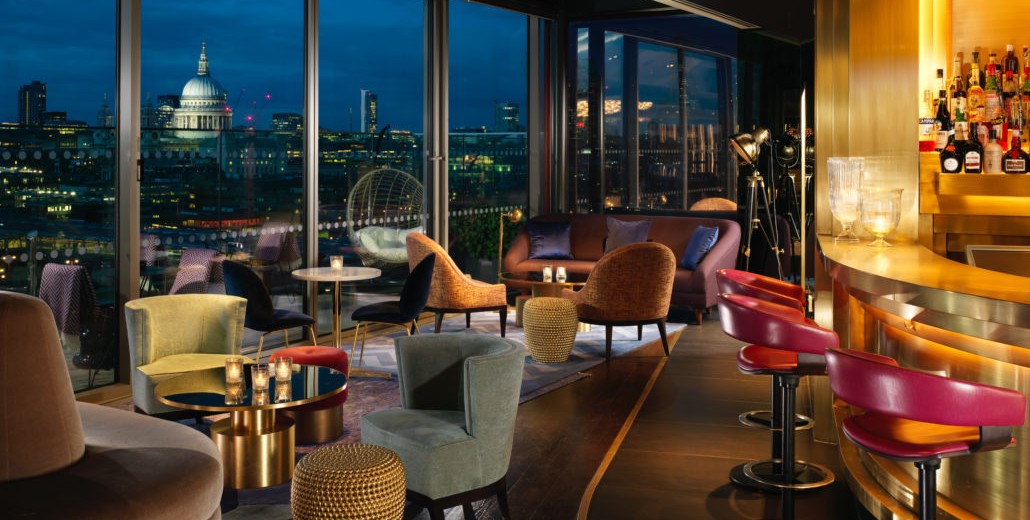 A chic rooftop lounge in London with vibrant seating and large windows showcasing a view of St. Paul’s Cathedral at night.
