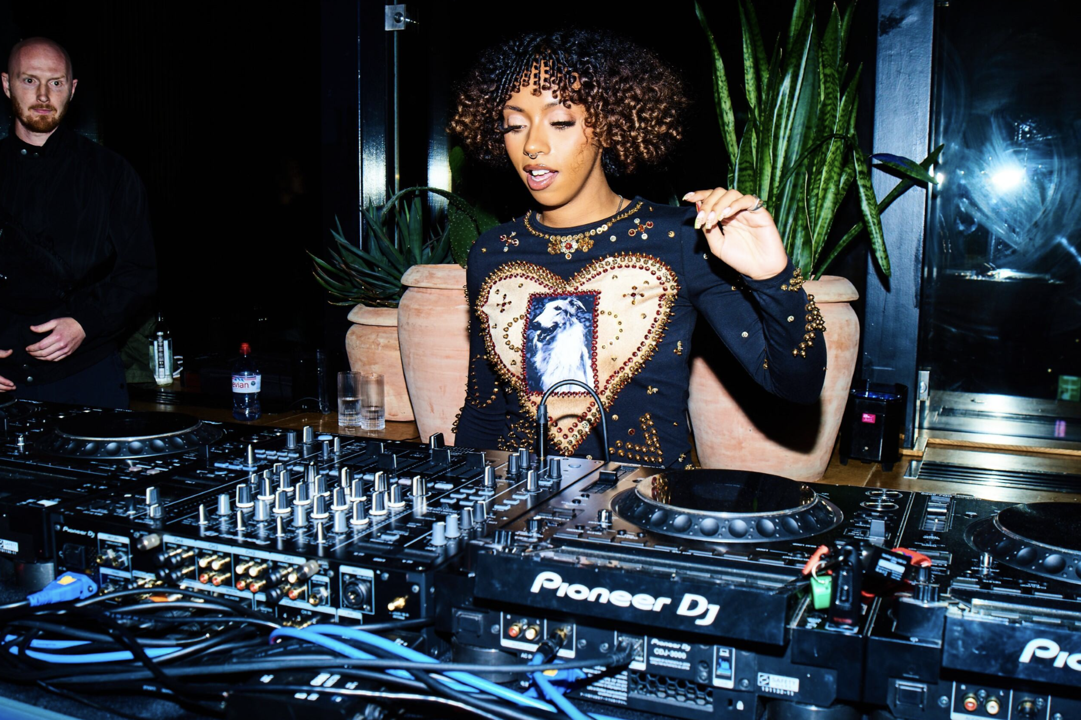 DJ performing at a Pioneer DJ setup, wearing a black top with gold accents and a heart design, plants in the background.