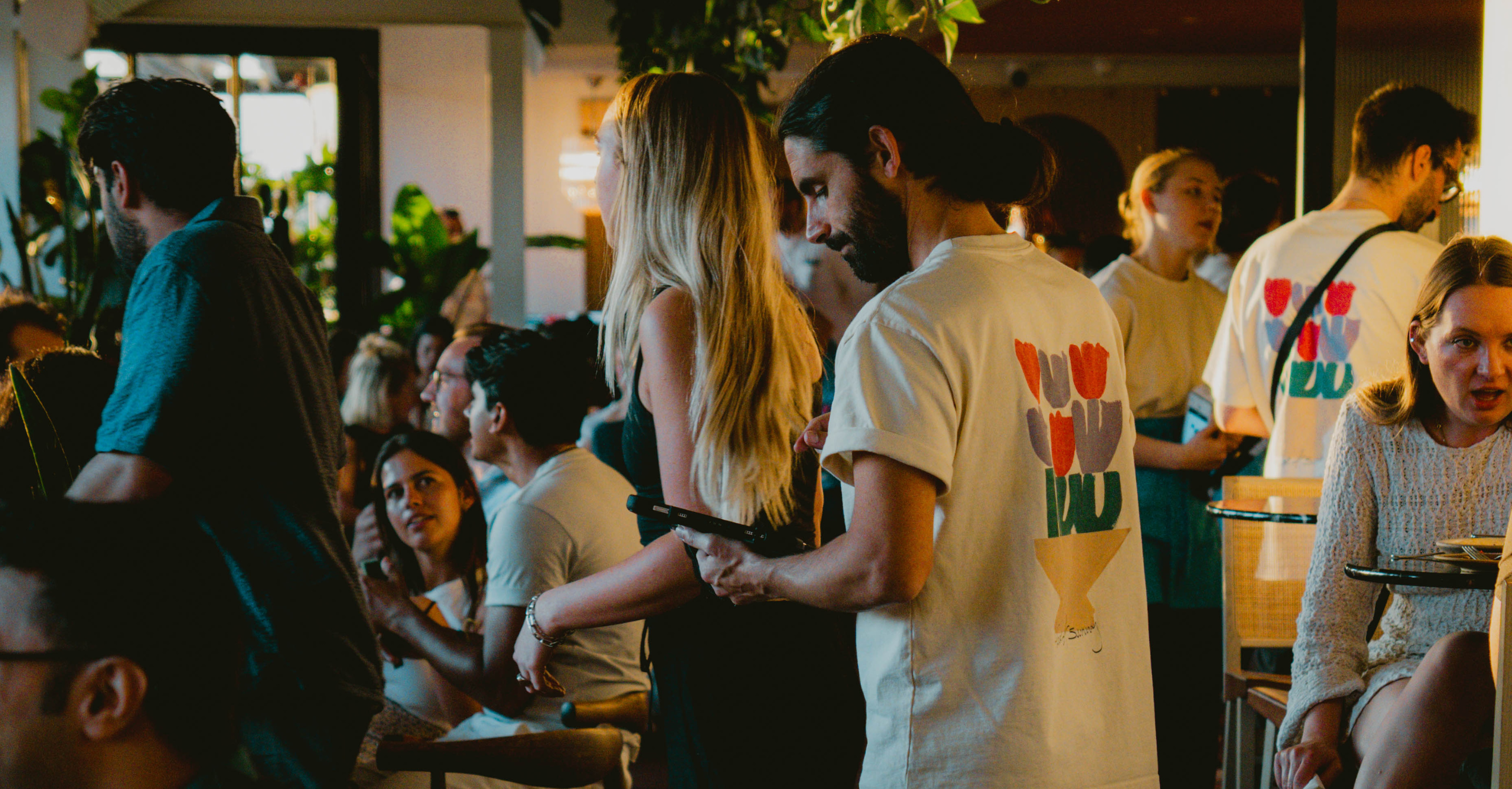 A lively bar scene where a male server in a floral-printed T-shirt in a dimly lit, stylish space filled with greenery.