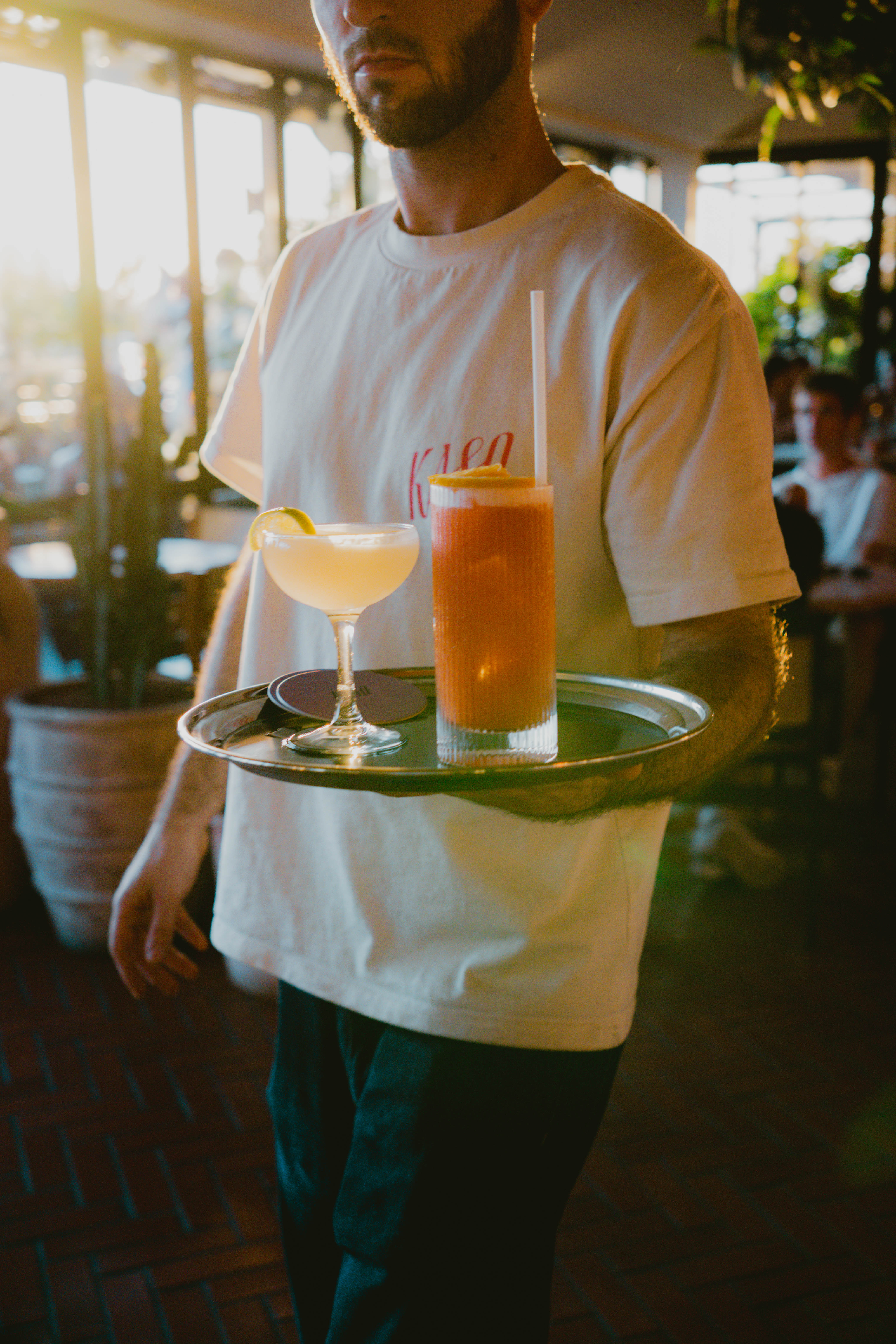 Bartender in a white t-shirt carries a tray with two colourful cocktails in a lively bar setting at Kaso.