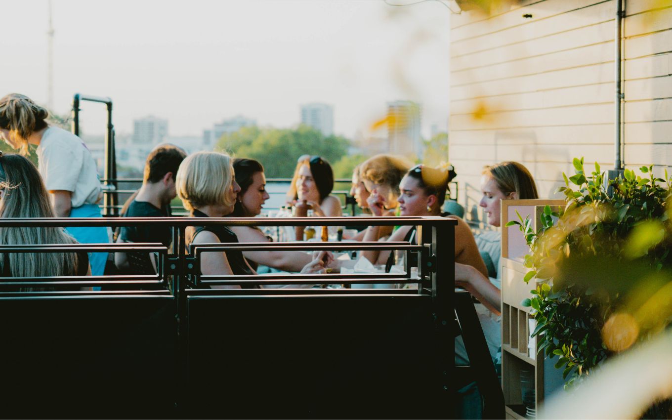 A group of people enjoying drinks and conversation on a rooftop terrace at sunset, with city buildings in the background.