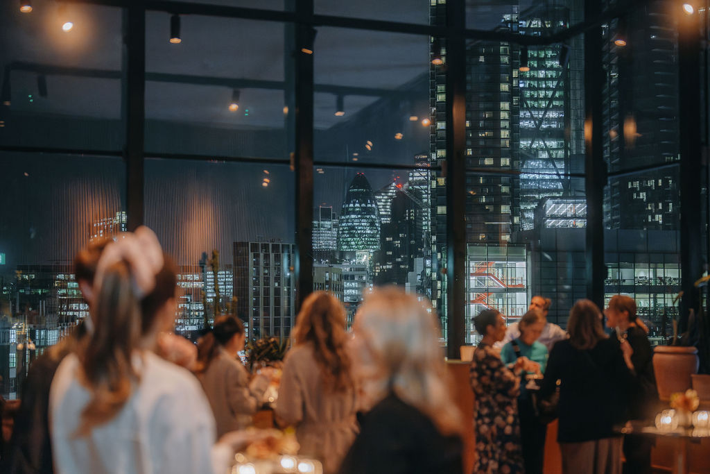 A bustling evening event with guests socializing in a lounge with large windows framing London’s cityscape at night.