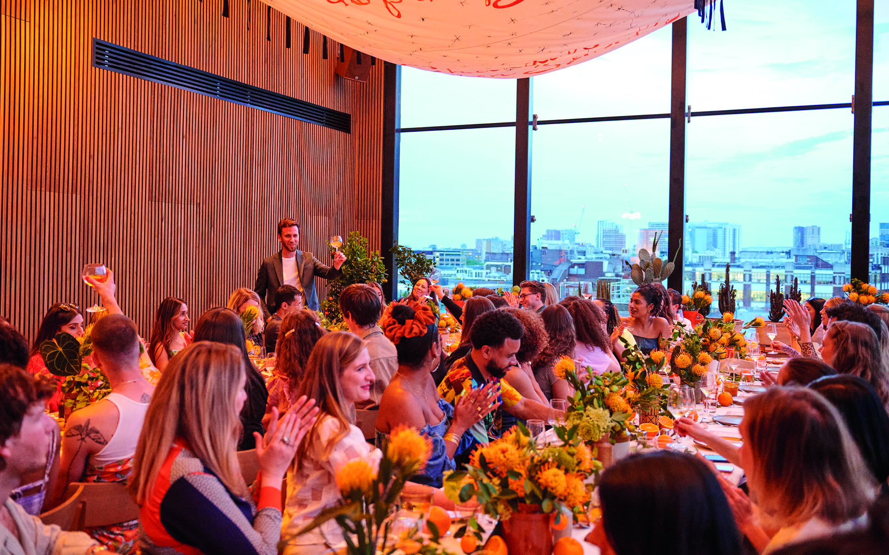 A lively dining event in London with guests seated at a long table adorned with bright yellow flowers and greenery.