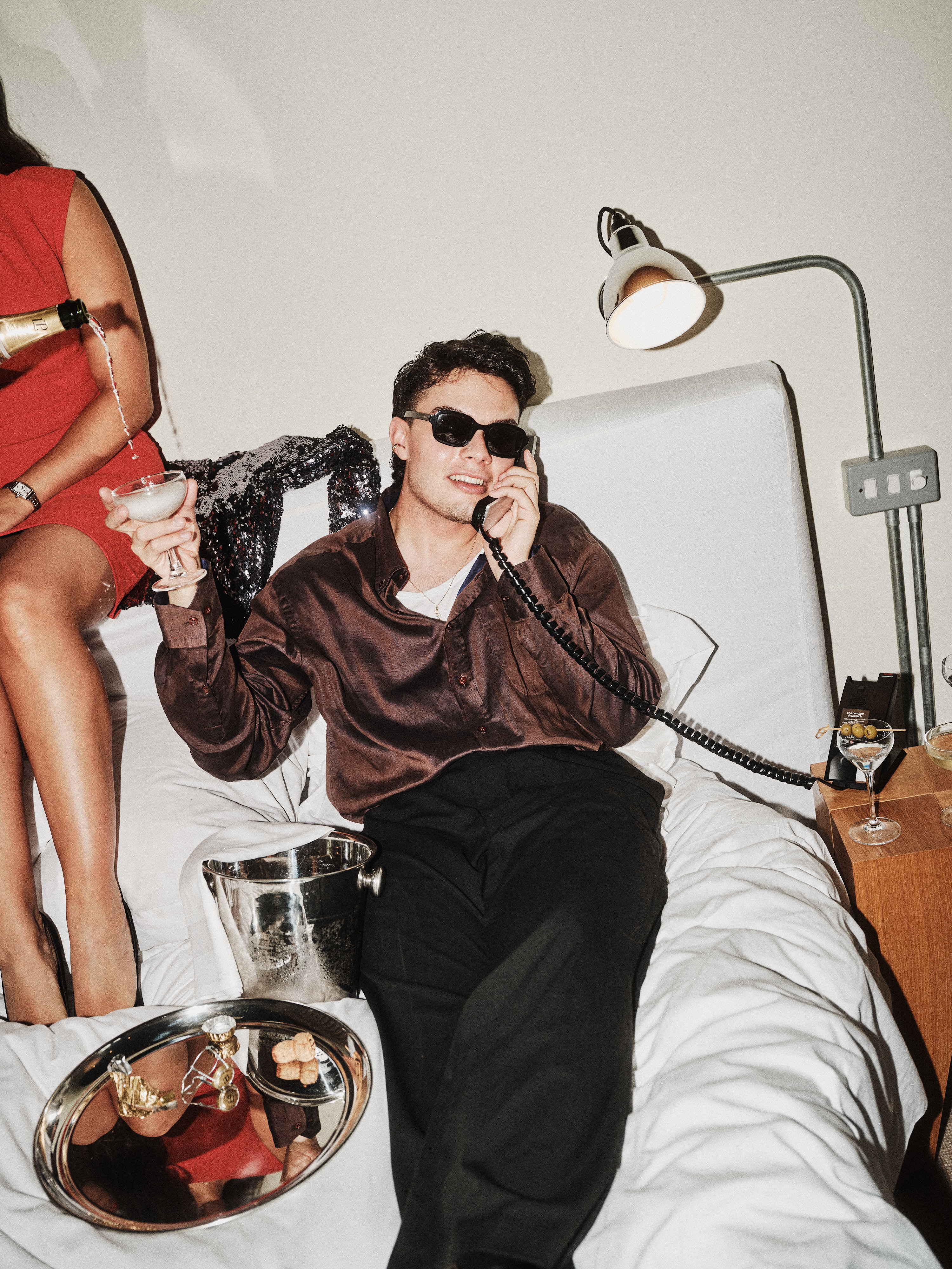 A man in sunglasses and a silk shirt lounging on a hotel bed, talking on a vintage phone and holding a cocktail.