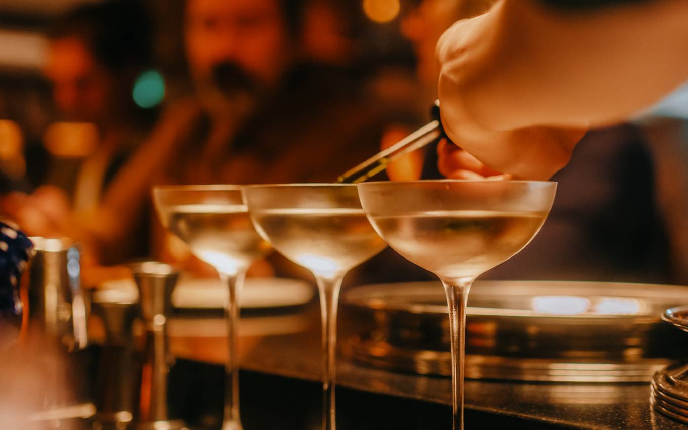 A bartender using tweezers to garnish three coupe glasses filled with clear cocktails in a dimly lit bar in London.