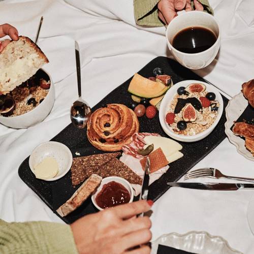 A breakfast spread on a bed, including fresh fruit, pastries, granola, cheese, bread, and coffee, with hands reaching in.
