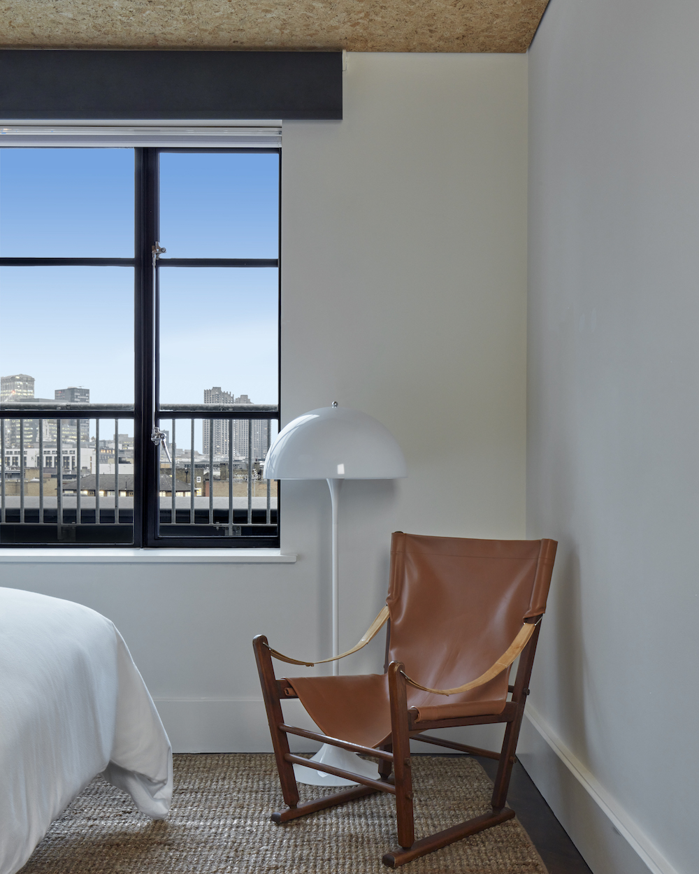 Contemporary hotel room in London with leather lounge chair, white floor lamp, and window view to the city.