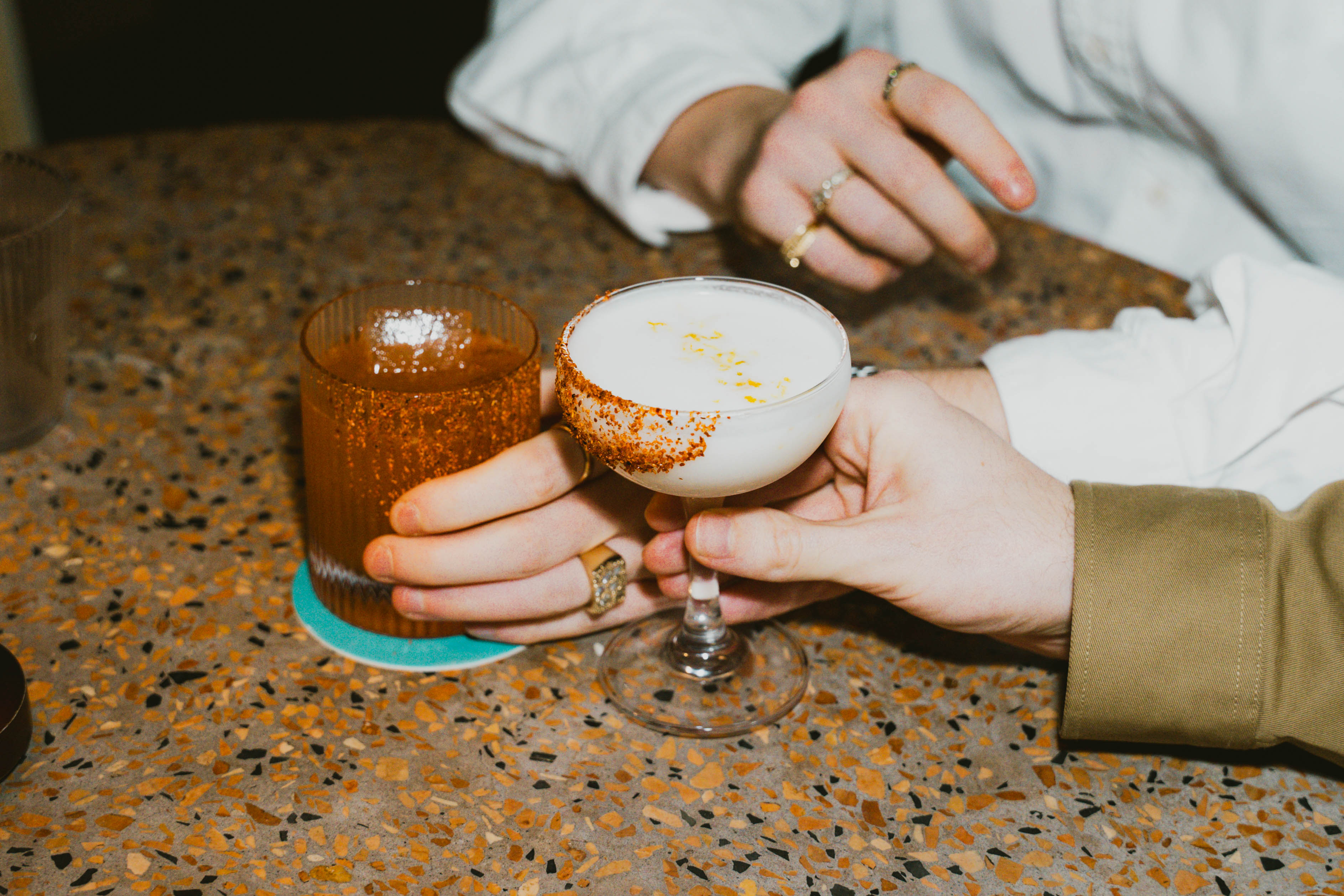 Two hands holding cocktails—one in a coupe glass with a chili rim, the other in a textured tumbler—on a speckled table.