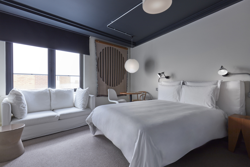 Modern hotel room in London with a white bed, sofa, circular light fixture, and a minimalist art tapestry on the wall.