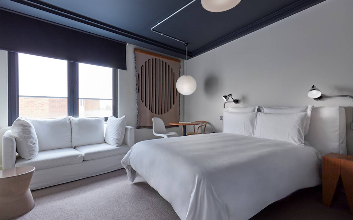 Modern hotel room in London with a white bed, sofa, circular light fixture, and a minimalist art tapestry on the wall.