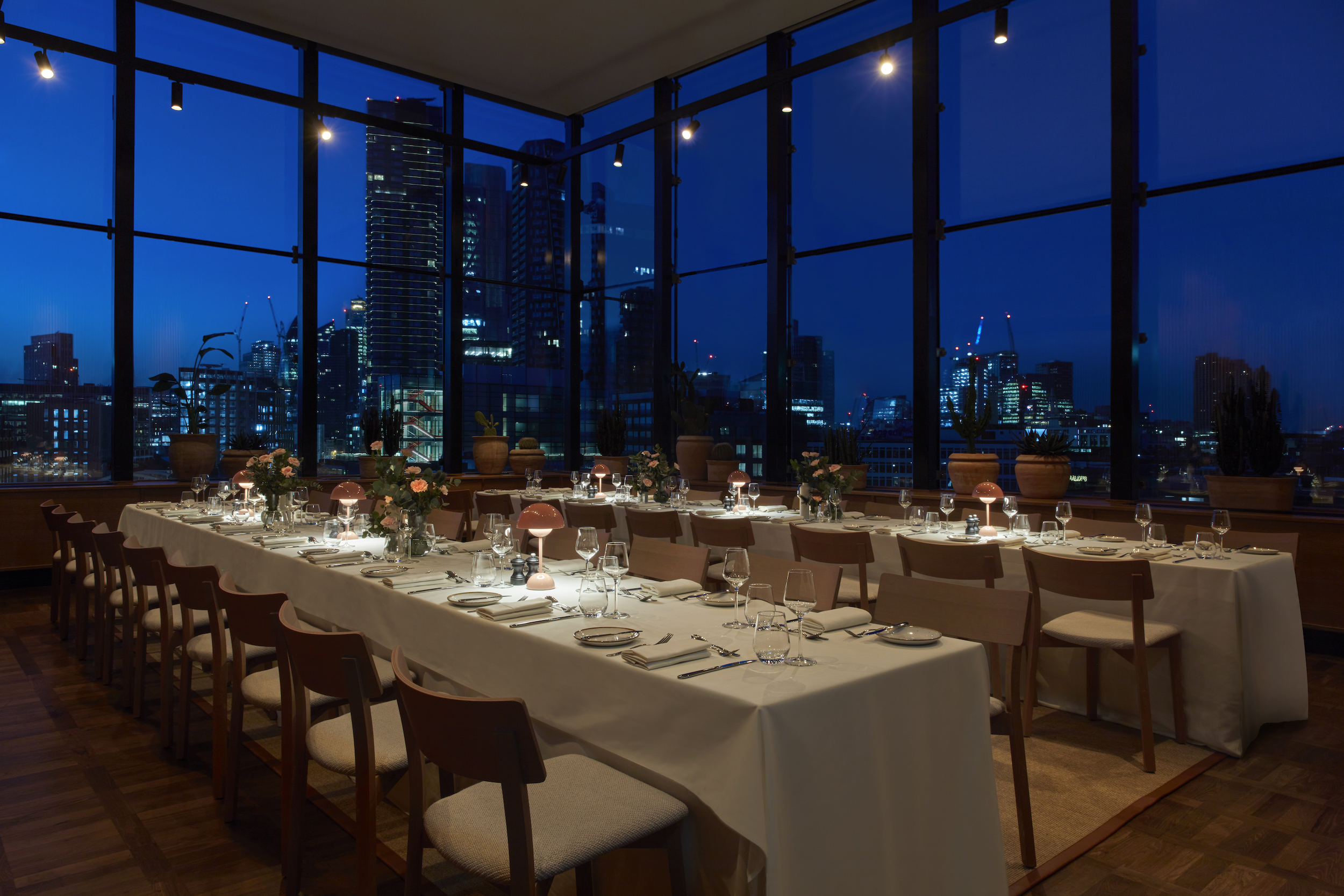 Elegant dining area with pink lamps and flowers on long tables, overlooking a city skyline through large windows at night.