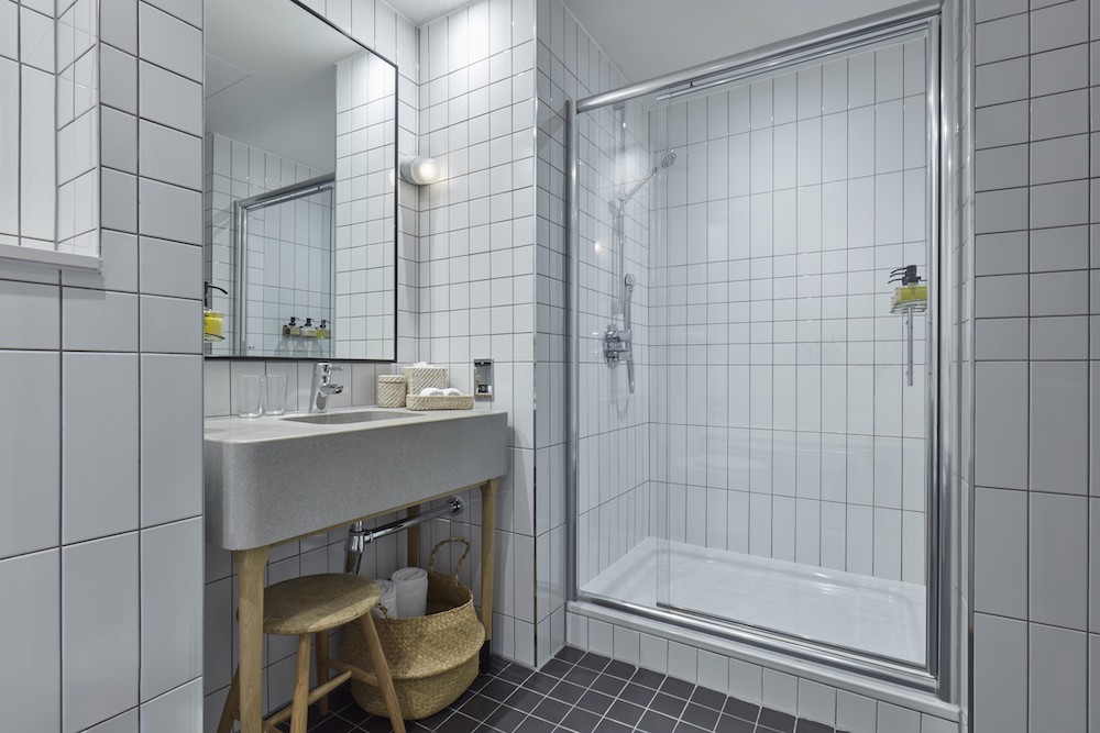 A sleek bathroom with white tiled walls, a glass-enclosed shower, a modern sink, and a wooden stool with a woven basket.