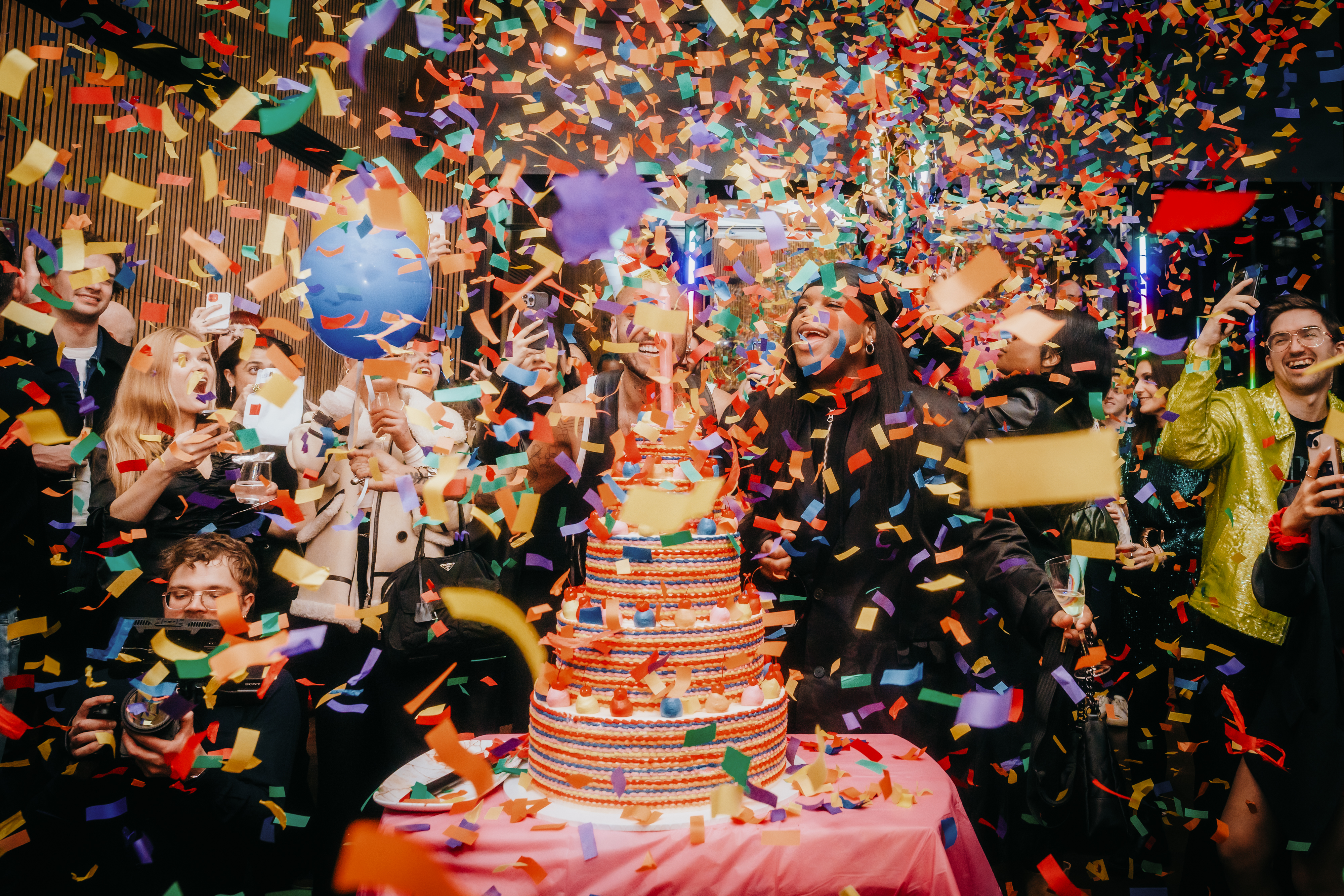 A joyful group of people celebrating a birthday party, surrounded by colourful confetti and festive decorations.