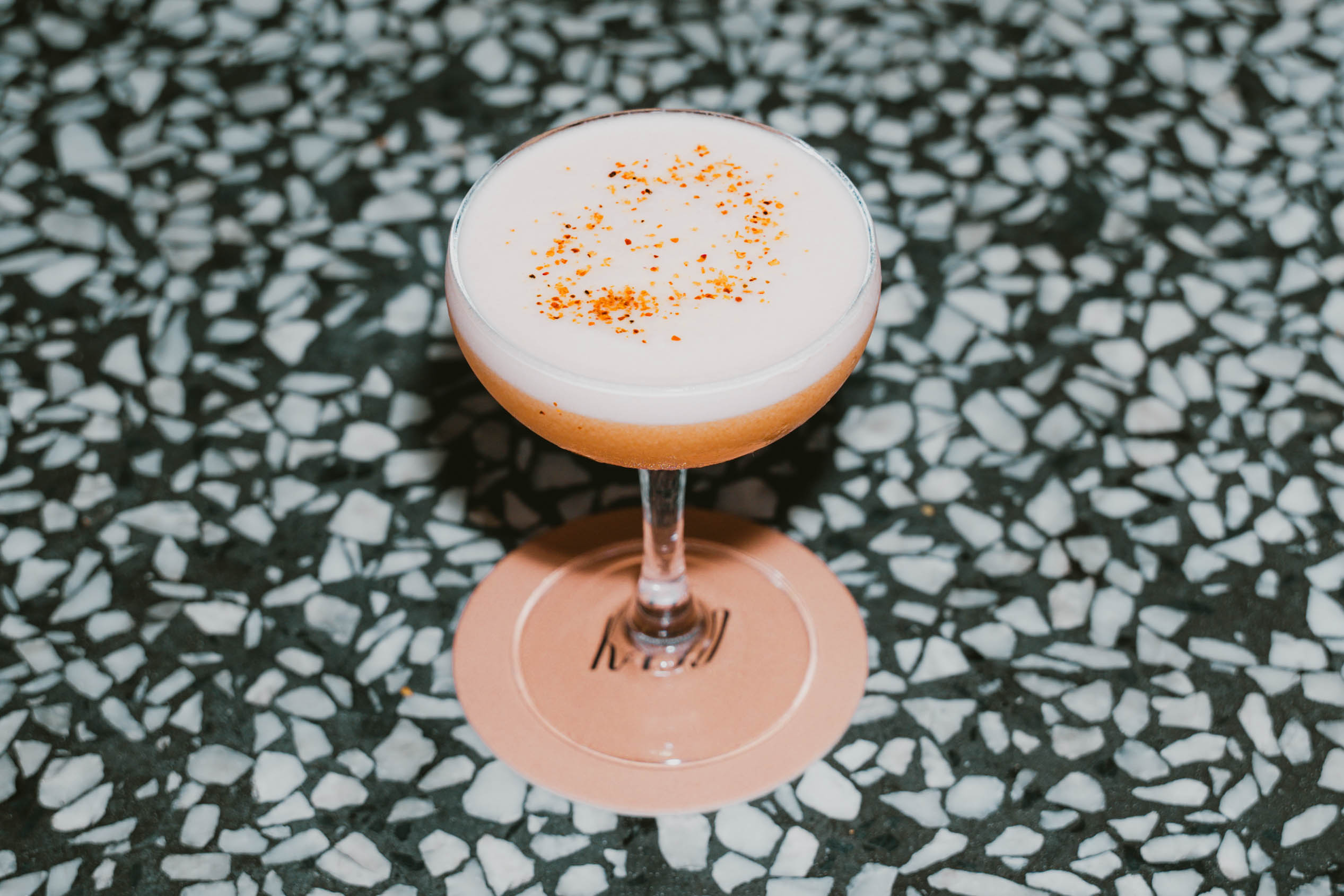 A frothy cocktail in a coupe glass, topped with a sprinkle of spice, sits on a terrazzo bar counter at Kaso.