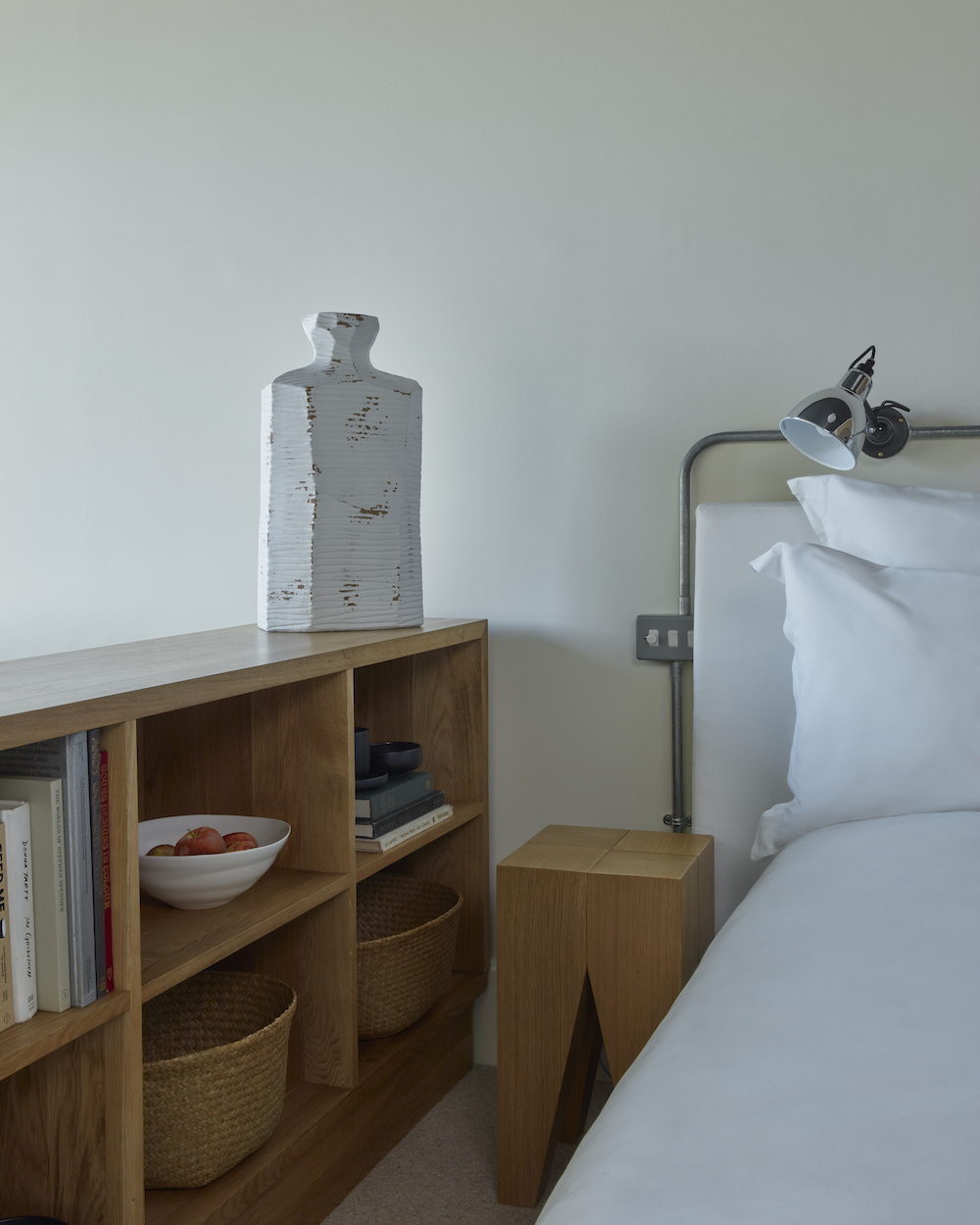 Minimalist bedside with white bedding, a metal reading light, and a wooden bookshelf with a vase in a hotel room in London.