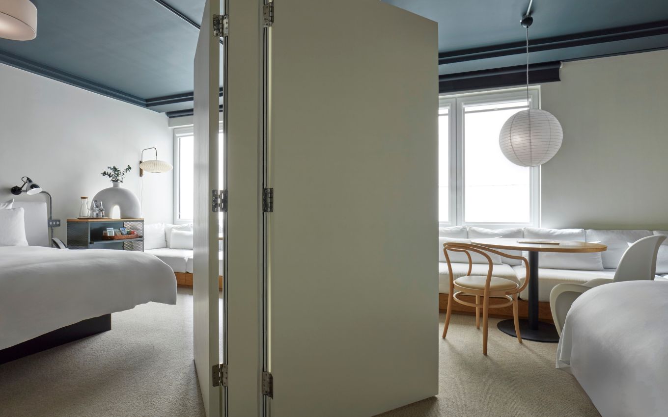 A spacious hotel room in London with a foldable partition, two beds, a seating area, and large windows letting in natural light.