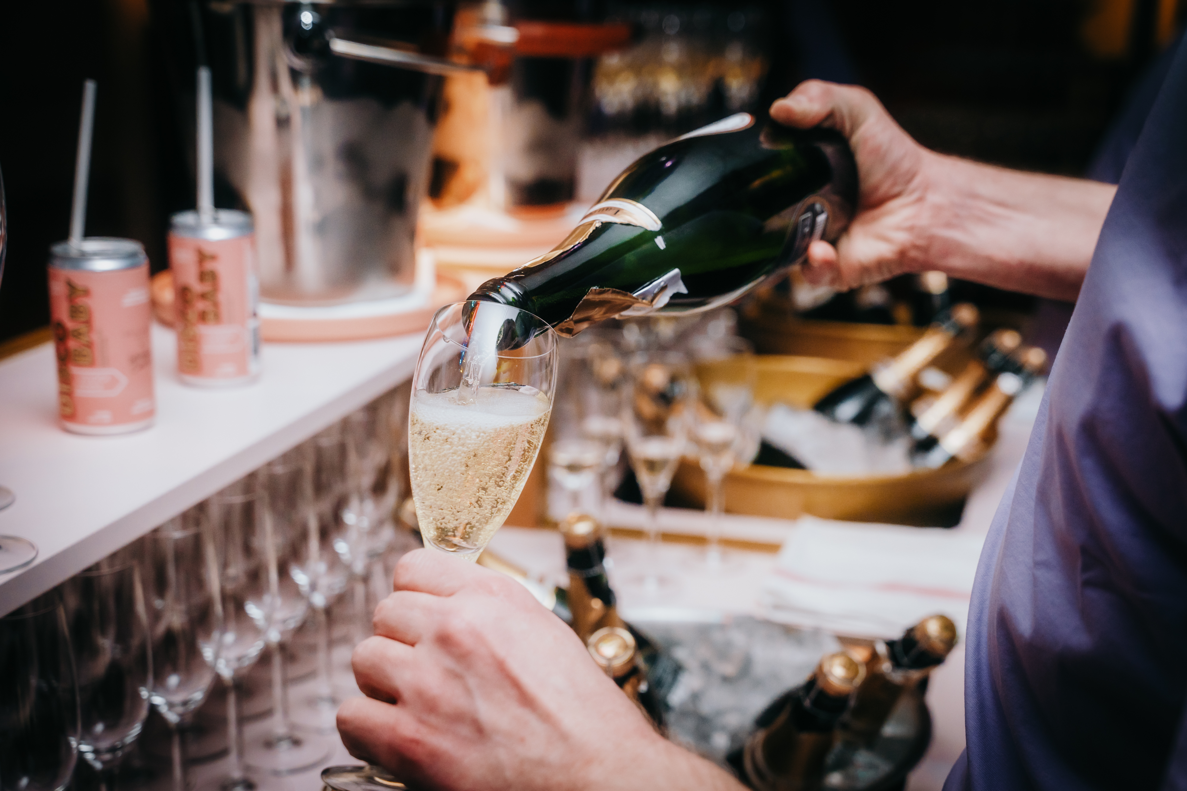 Person pouring sparkling wine into a glass at a bar setup with rows of glasses, cans, and ice buckets.