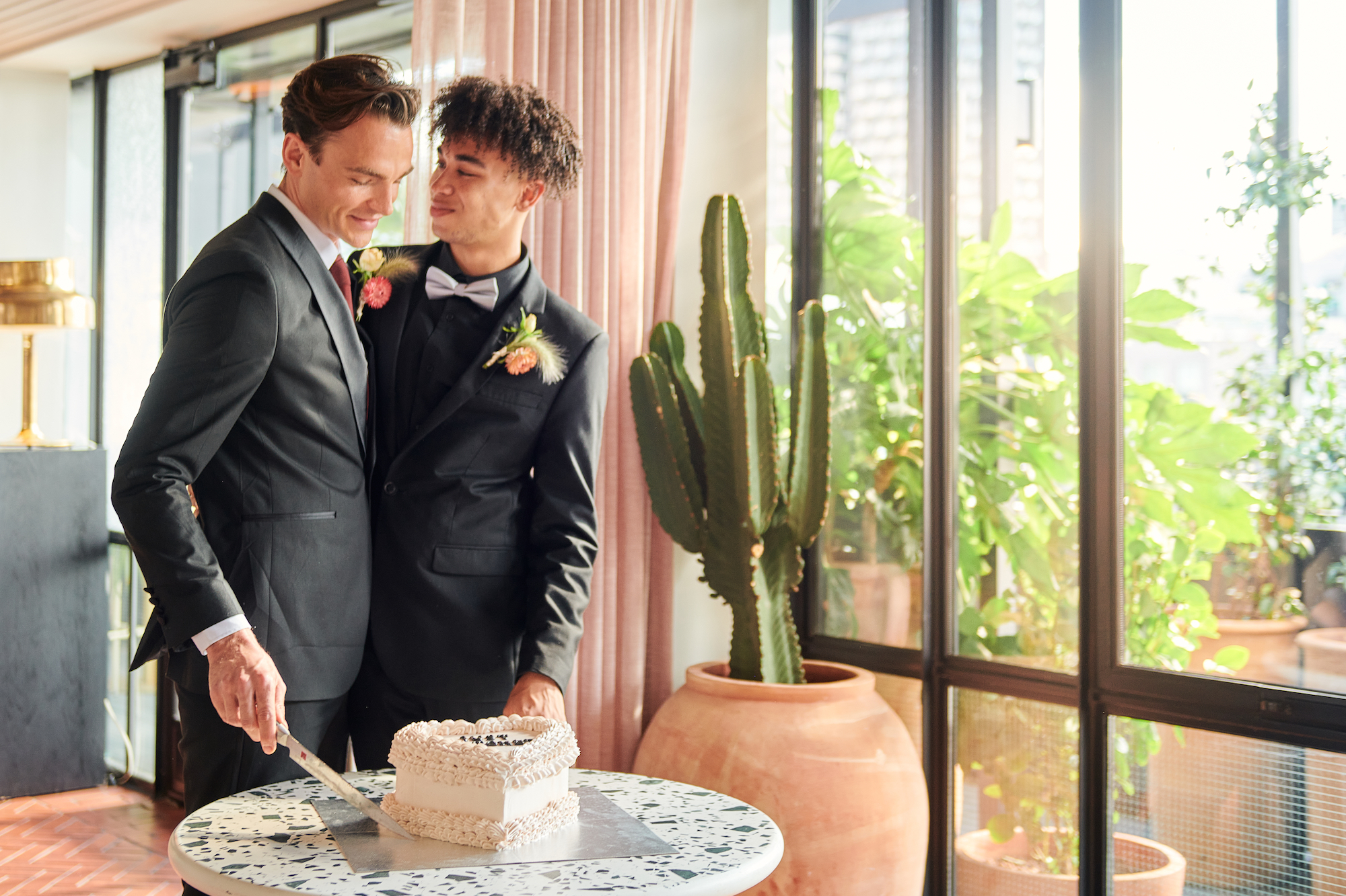 Two men in suits sharing a wedding moment while cutting a cake, standing beside large potted plants and glass windows.