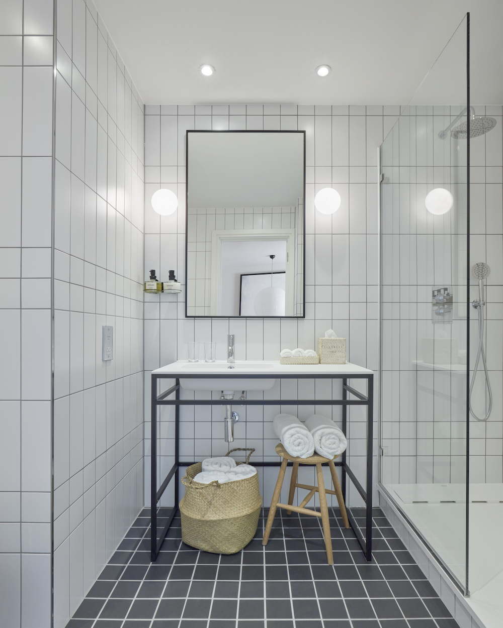White-tiled hotel bathroom with a minimalist sink, rolled towels, and woven baskets on the black and white tiled floor.