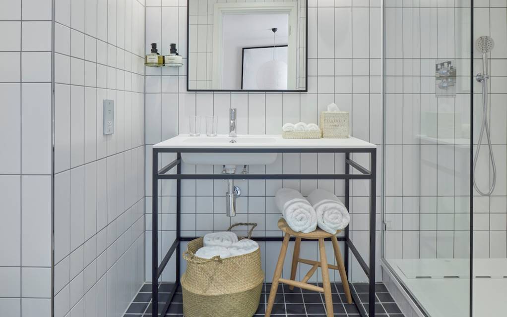 White-tiled hotel bathroom with a minimalist sink, rolled towels, and woven baskets on the black and white tiled floor.