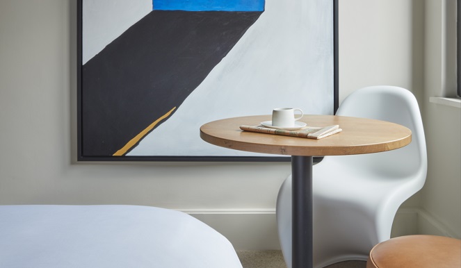 Modern art piece with bold blue and black shapes, hung beside a small wooden table and a white chair at a London hotel room.