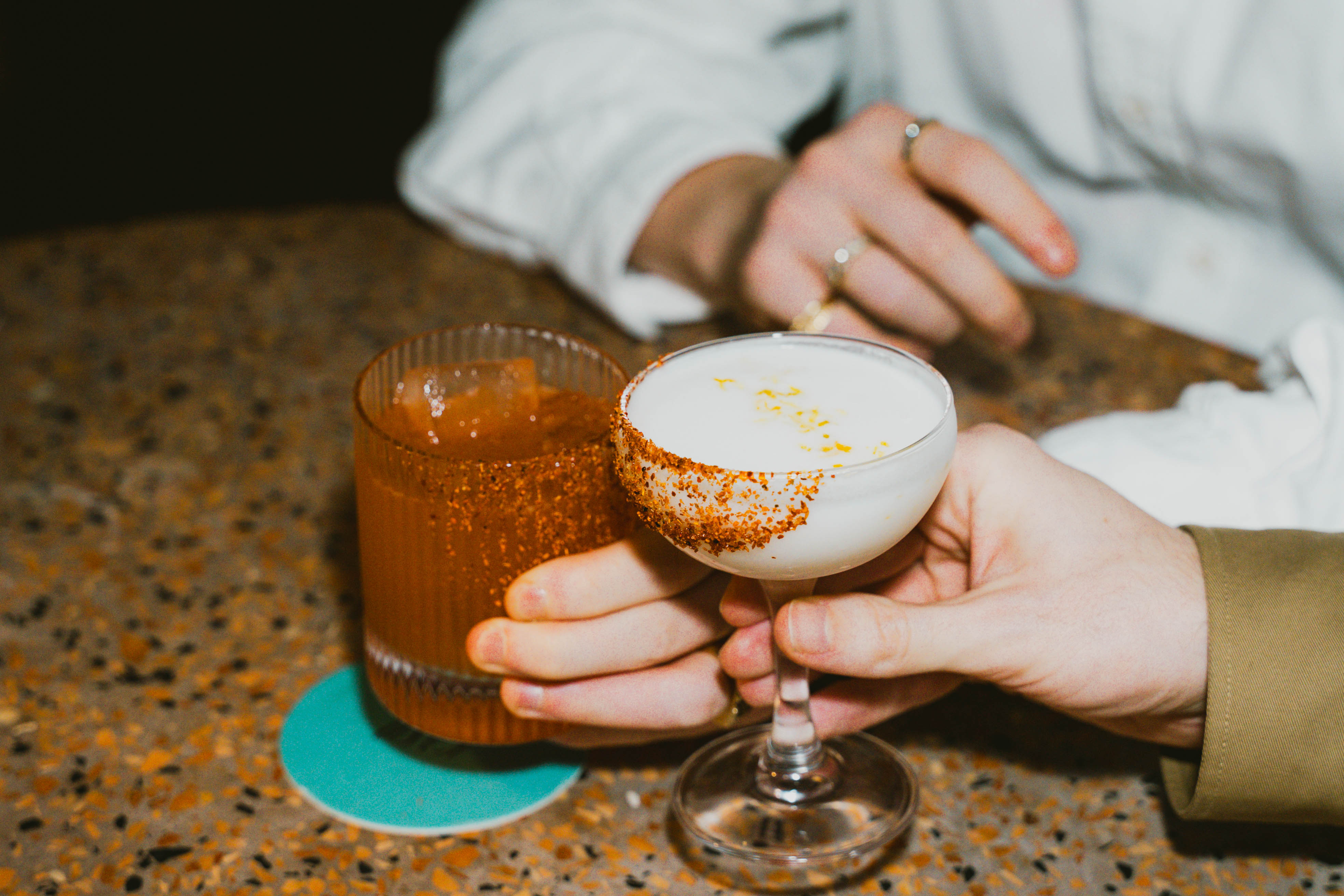 Two hands holding cocktails, one with a salted rim and frothy top, the other a spicy margarita with a chilli-rimmed glass.