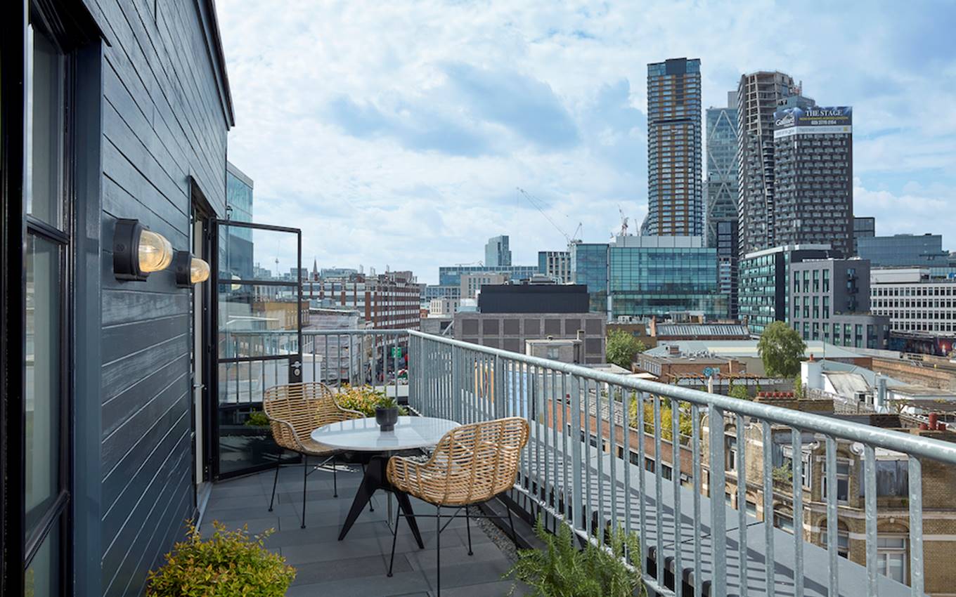 Rooftop balcony with rattan chairs and table, with London skyline views on a clear day, showing a relaxing urban retreat.