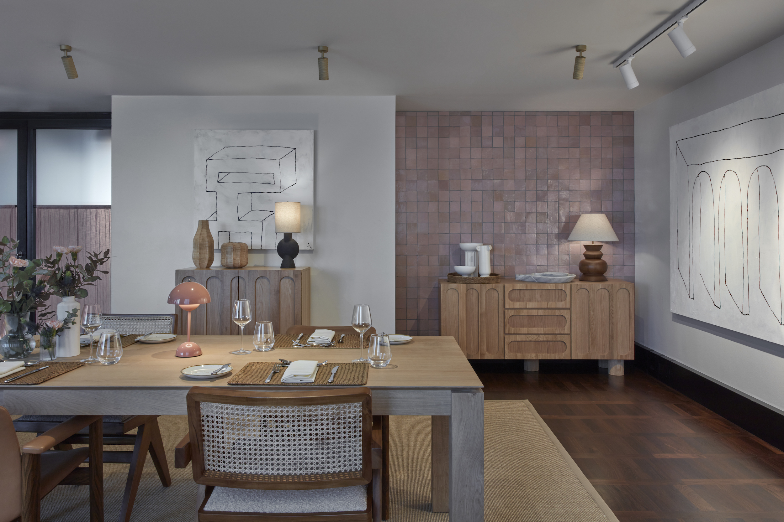 Dining area with wooden table, pink wall tiles, artistic decor, and place settings for guests, creating a cosy ambiance.
