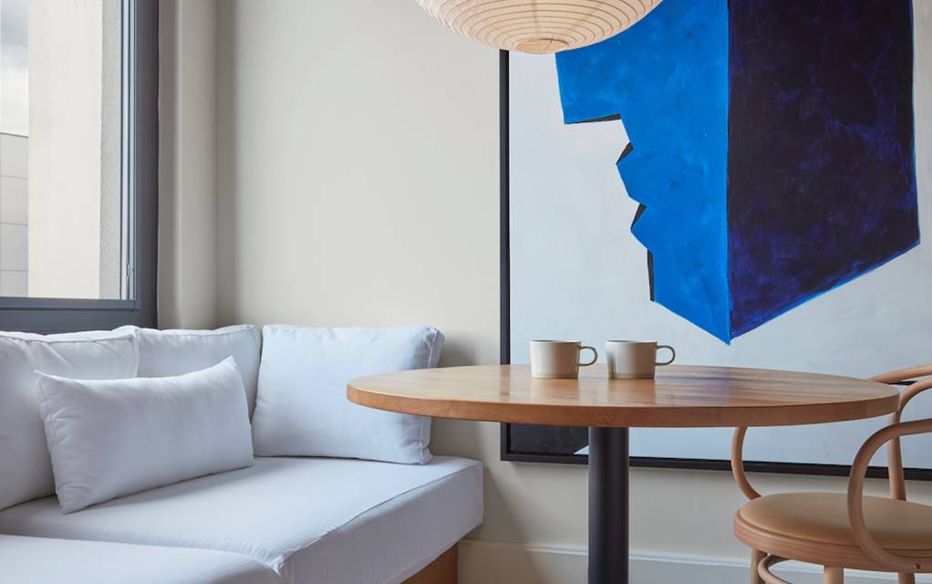 Cosy hotel room corner with a round wooden table, two cups, and a large blue abstract painting beside a soft white bench.