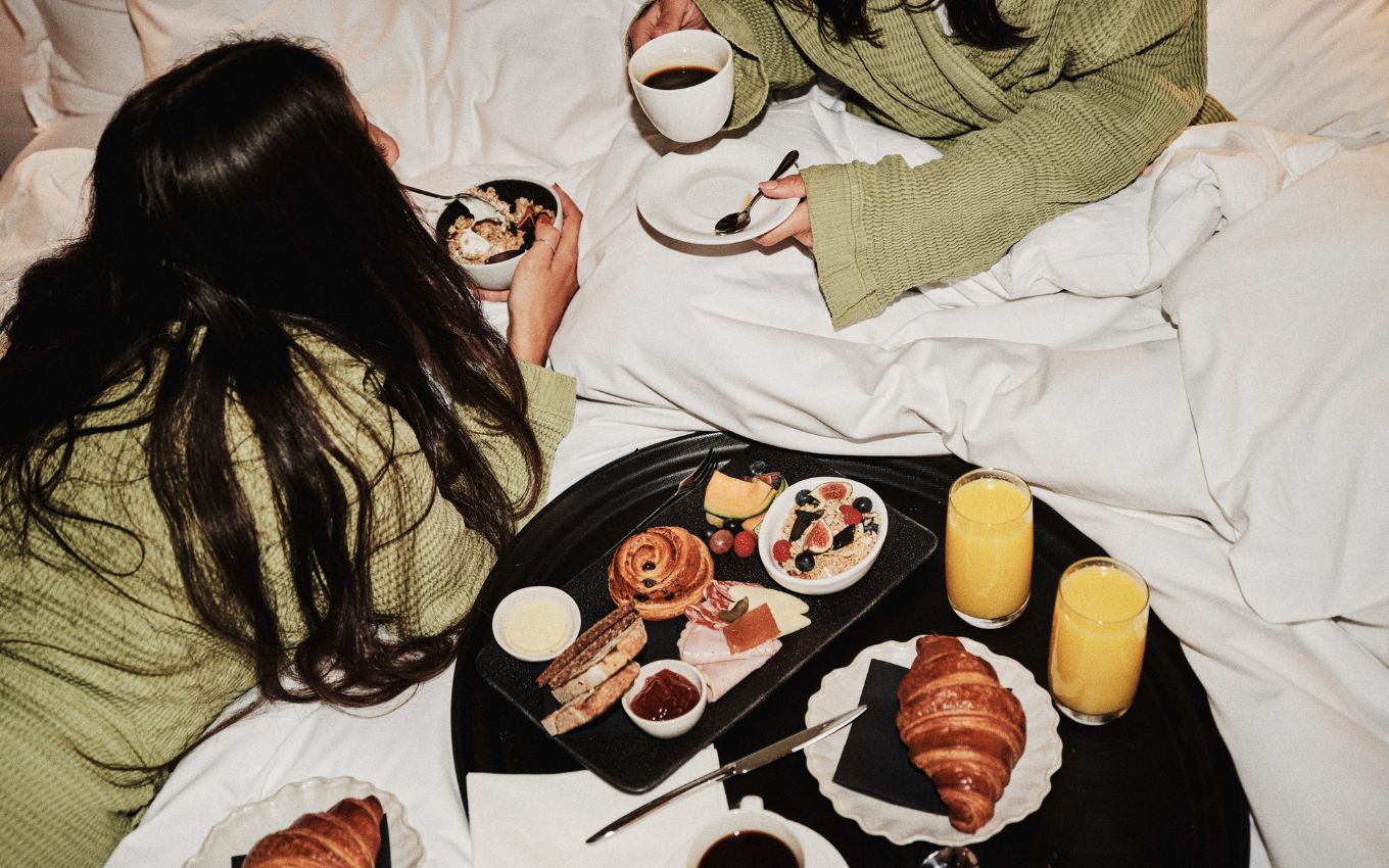 Two women in green robes enjoying a breakfast in bed with croissants, granola, fresh fruit, and glasses of orange juice.