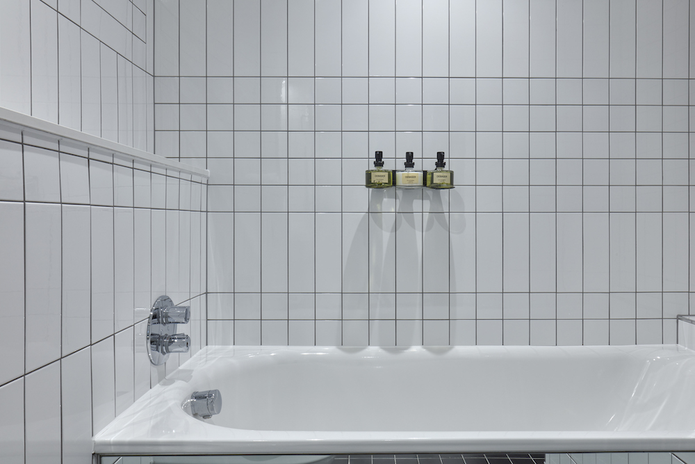 Modern white tiled bathtub with mounted bath products against the wall in a hotel room at One Hundred Shoreditch in London.