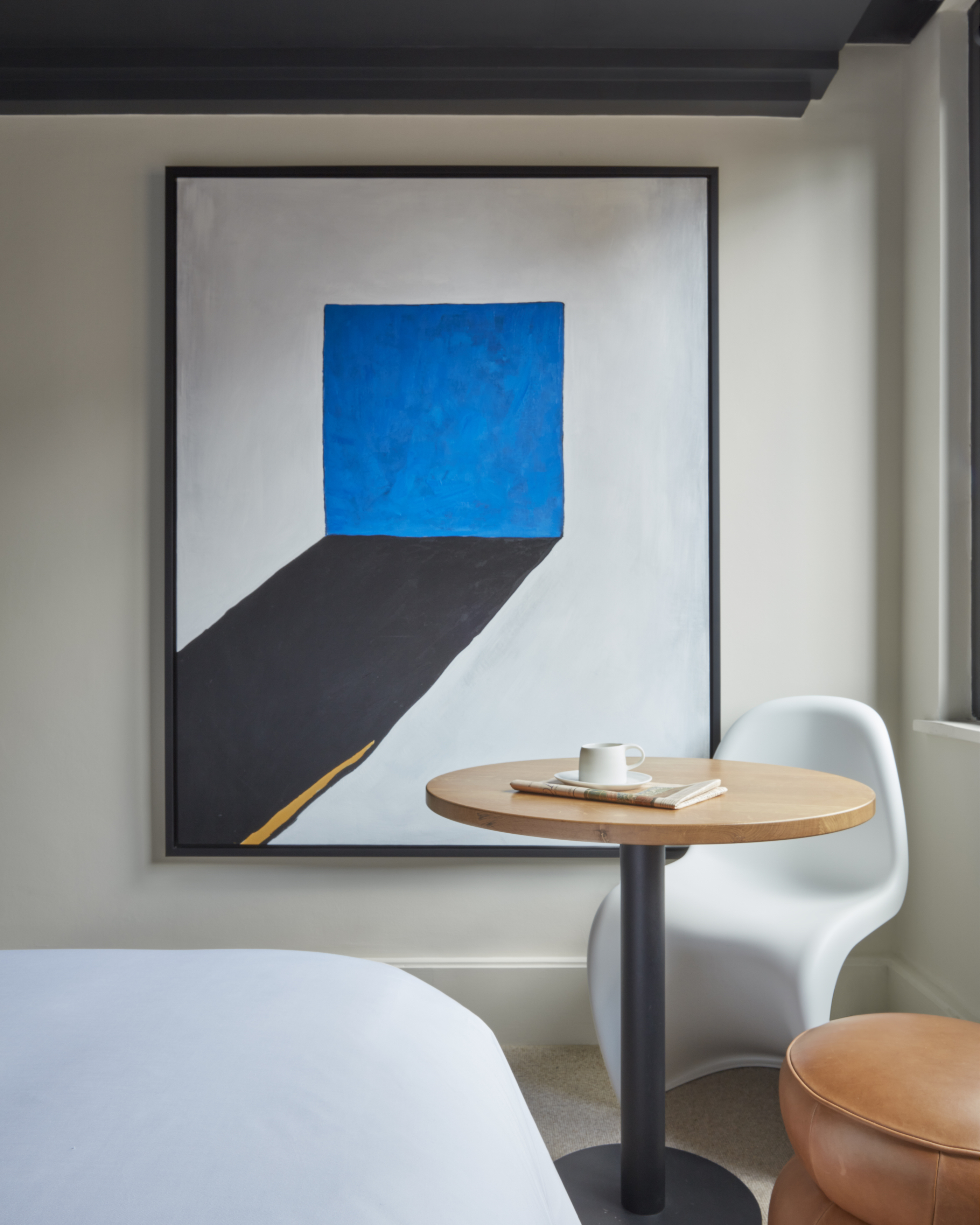 Modern art piece with bold blue and black shapes, hung beside a small wooden table and a white chair at a London hotel room.
