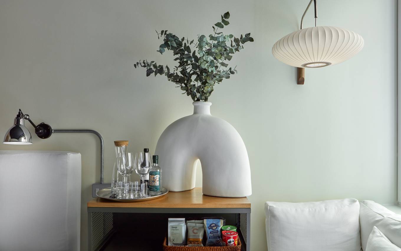 Vase with eucalyptus branches beside glassware and snacks on a wooden side table in a cozy, in a minimalist hotel room.